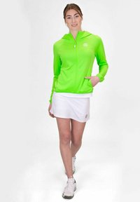 Felpa verde brillante con zip e cappuccio, caratterizzata da una tasca frontale. Abbinata a una gonna bianca e a scarpe sportive bianche.