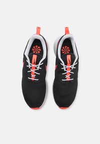 Sapatos desportivos em malha preta com detalhes em vermelho e solas brancas. Inclui um logo em forma de flor e um design de atacadores tradicional.