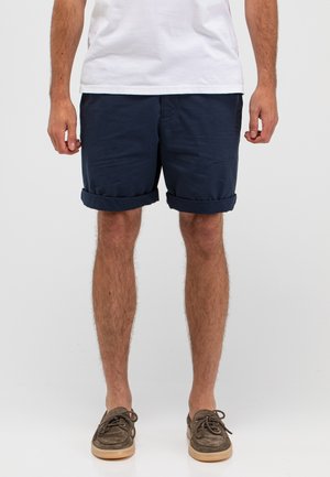 Mann trägt marineblaue, hochgekrempelte Shorts, ein weißes T-Shirt und braune Freizeitschuhe, steht vor einem schlichten hellen Hintergrund.