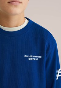 Modra teksturirana pulover s kapuco v modri barvi, belim ovratnikom in logotipom "BLUE RIDGE DENIM" na prsih. Ima rebrast dizajn.