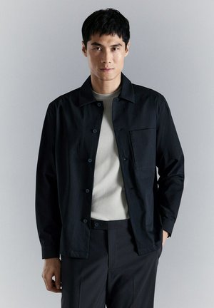 Uomo che indossa una giacca nera con bottoni sopra una camicia grigio chiaro, con pantaloni neri, in piedi davanti a uno sfondo grigio uniforme.