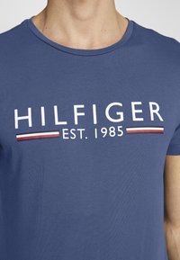 T-shirt di cotone blu con collo a giro, maniche corte e il testo "HILFIGER EST. 1985" in bianco, con accenti a righe rosse e bianche.