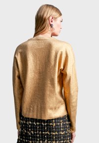 Guld, texturerad tröja med avslappnad passform och långa ärmar, i kombination med en svart rutig kjol med metalliska detaljer.