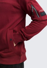 Felpa bordeaux con finitura testurizzata, dotata di tasca laterale con zip e maniche a coste. Include una toppa con logo sul braccio.