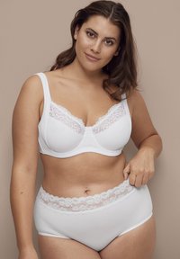 Vrouw met een gemiddelde huidtint en bruin haar die een witte bh met kant en hoge onderbroek draagt, en voorzichtig de tailleband vasthoudt.