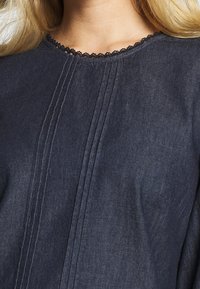 Top in denim scuro con scollatura in pizzo nero a scallop, pieghe verticali sul centro davanti e una finitura morbida e testurizzata.