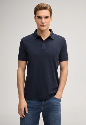 Joven con cabello corto castaño claro que lleva una camiseta tipo polo azul marino oscura y vaqueros azules, de pie con una mano en el bolsillo.