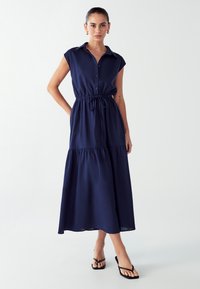 Jūrų laivyno mygtukų suknelė su trumpomis rankovėmis, su raišteliu juosmenyje ir sluoksniuota sijonu. Lygus audinys, kulkšnies ilgio, derinama su juodomis šlepetėmis.