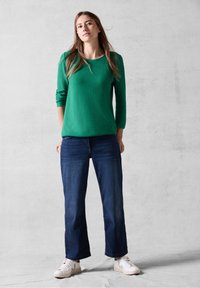 Maglia verde a maniche lunghe con motivo testurizzato, abbinata a jeans blu scuro e sneakers bianche. La vestibilità è rilassata e informale.