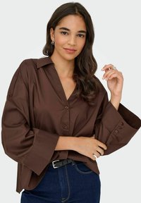 Jeune femme aux longs cheveux noirs portant une chemise marron ample à boutons et un jean foncé, posant avec une main près du visage.