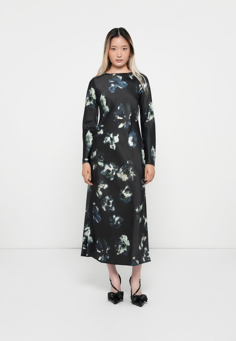 Long black floral dress with green and blue abstract patterns. Smooth texture, long sleeves, round neckline, paired with black bow sandals.