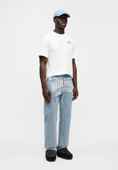 Λευκό T-shirt με μπλε λογότυπο, ανοιχτό μπλε τζιν παντελόνια με έντονο σχέδιο, σκούρες παντόφλες με φλοράλ σχέδιο και ένα καπέλο που ταιριάζει.