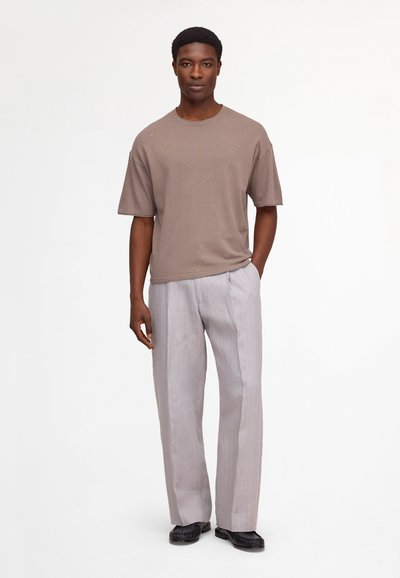 Homme debout, main dans la poche, portant une chemise marron ample à manches courtes, un pantalon large gris clair et des chaussures noires sur fond blanc.