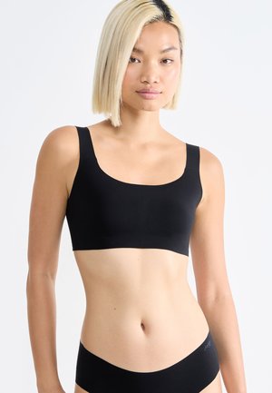 Frau mit kurzen blonden Haaren, die einen schwarzen Sport-BH und passende nahtlose schwarze Unterwäsche vor weißem Hintergrund trägt.