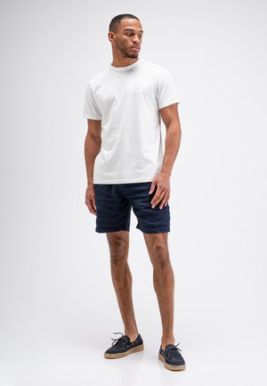 Man die staat met een witte korte mouwen T-shirt, donkerblauwe korte broek en zwarte casual schoenen tegen een effen lichte achtergrond.