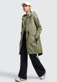 Persoană tânără îmbrăcată cu un parka cu glugă de culoare verde deschis, pantaloni cargo negri, pantofi sport albi și o șapcă de baseball albă, stand pe un fundal neutru.