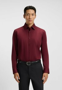 Camicia a maniche lunghe borgogna con una texture liscia, davanti con bottoni e colletto classico, abbinata a pantaloni eleganti neri e cintura.