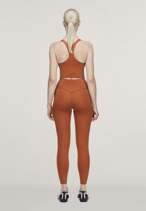 Une femme se tient de dos, présentant un soutien-gorge de sport et un legging couleur rouille avec des baskets noires et blanches, sur un fond de studio uni.