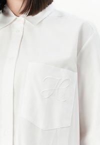 Woman wearing a white button-up shirt with a chest pocket embroidered with the letter H and "HUGO" text in white thread.