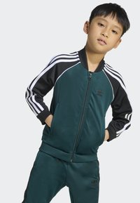 Mörk teal träningsdräkt med svarta ärmar, vitrandig design, zip-up jacka och ribbade ärmslut. Har Adidas-logotyp på bröstet och byxorna.