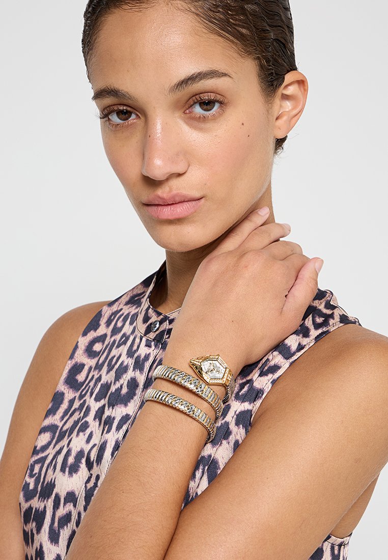 Orologio esagonale dorato con bracciali testurizzati in argento e oro, abbinato a un top senza maniche con stampa leopardo. Pelle liscia, sfondo neutro.
