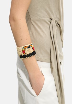 Ręka z dłonią w białej kieszeni spodni nosząca szeroką beżową bransoletkę ozdobioną kolorowymi koralikami i czarnym zygzakowatym wzorem.