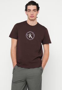 Pruun puuvillane t-särk, millel on ümmargune logo valge tekstiga "Calvin Klein New York 1968" ja "CK" keskel. Lühikesed varrukad, ümarselg.