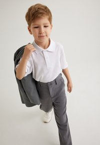 Camisa polo blanca con tres botones, pantalones de vestir grises con bolsillos laterales y una chaqueta gris colgada de un brazo. Zapatillas blancas.