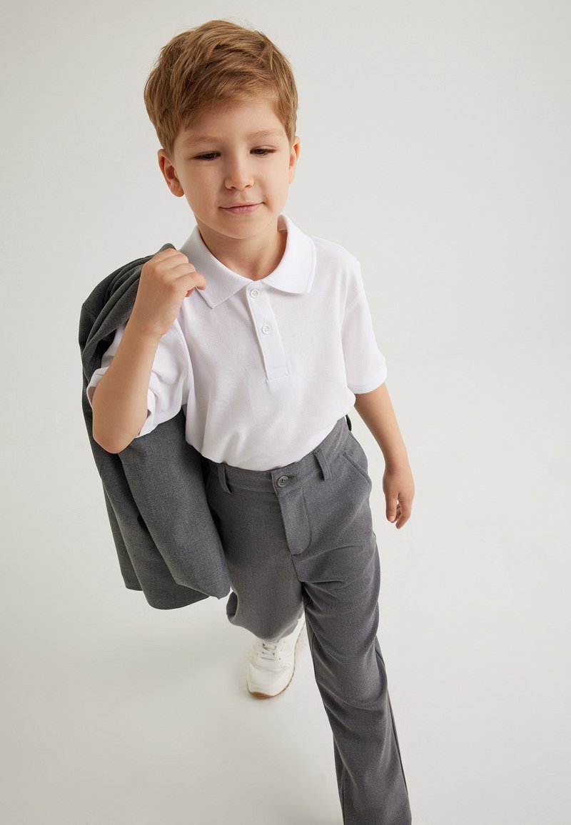 Camisa polo blanca con tres botones, pantalones de vestir grises con bolsillos laterales y una chaqueta gris colgada de un brazo. Zapatillas blancas.