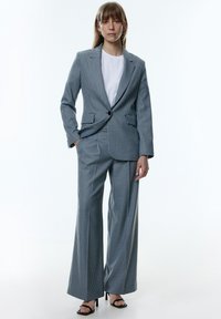 Gestreepte pakken in licht en donkerblauw stof, met een blazer met één knoop, brede broekspijpen en een eenvoudig wit topje eronder. Zwarte hoge hakken.