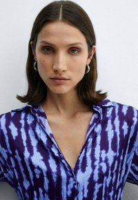 Vrouw met steil bruin haar, gekleed in zilveren oorbellen en een blauwe en paarse blouse met patroon, tegen een effen licht achtergrond.