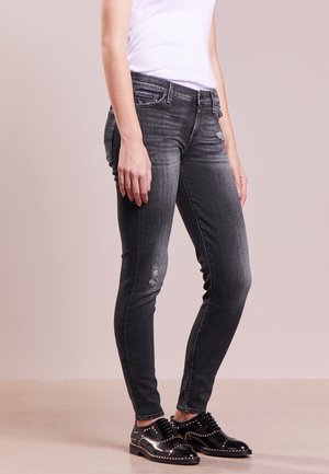 Person iført falmede sorte skinny jeans med små slidte lapper og sorte oxfordsko med nitter, stående på et glat gulv.