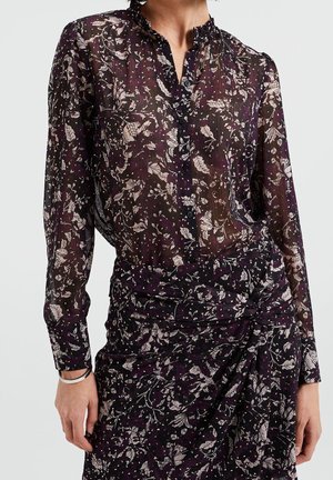 Gennemsigtig sort bluse med lange ærmer med hvid blomsterprint, parret med matchende højtaljede blomstrede omslagsnederdel, iført sølvarmbånd.