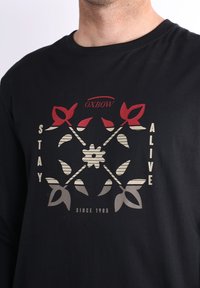 T-shirt à manches longues noir avec un imprimé graphique aux éléments rouges et beiges. Le texte indique « RESTEZ EN VIE » et « DEPUIS 1985 ».
