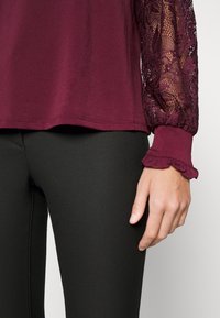 Burgunderfarbener Oberteil mit strukturiertem Körper und durchsichtigen Spitzenärmeln. Rüschenmanschetten sorgen für Detail. Kombiniert mit schwarzen, figurbetonten Leggings.
