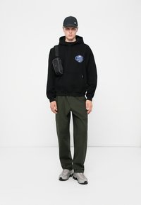 Zwarte hoodie met een voorzak en blauwe grafische afbeelding, gecombineerd met olijfgroene cargobroek. Grijze sneakers en een zwarte pet maken de outfit af.