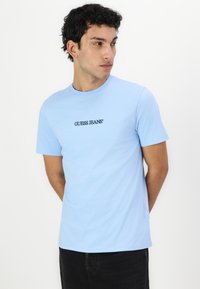 Heledsinine puuvillane T-särk lühikeste varrukatega, ringikujulise kaelusega, millel on must "GUESS JEANS" logo rinna peal.