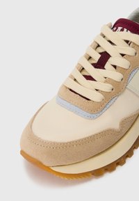 Primo piano di una sneaker beige e bordeaux con lacci crema e suola in gomma su sfondo bianco.