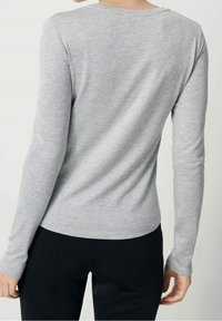 Mujer vestida con una camiseta de manga larga ajustada de color gris claro y pantalones negros, vista desde atrás contra un fondo simple.