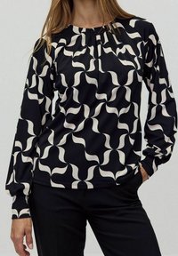 Langärmlige Bluse in Schwarz mit einem cremefarbenen, wellenartigen Muster. Mit einem gerafften Ausschnitt und einem glatten, leichten Stoff. Kombiniert mit schwarzen Hosen.