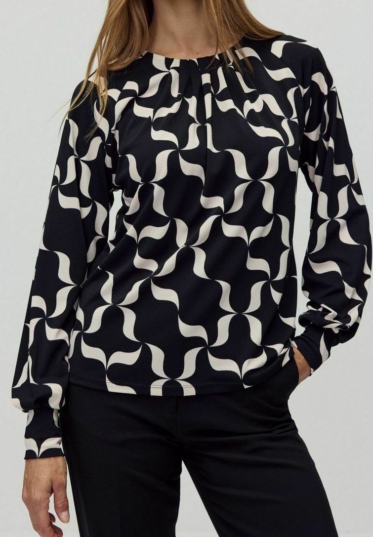 Langärmlige Bluse in Schwarz mit einem cremefarbenen, wellenartigen Muster. Mit einem gerafften Ausschnitt und einem glatten, leichten Stoff. Kombiniert mit schwarzen Hosen.