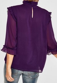 Blusa trasparente viola con scollo e maniche a rouches. La parte posteriore presenta una piccola apertura e un dettaglio a occhio di chiave, abbinata a jeans casual.