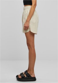 Bézs len rövidnadrág oldalsó hasítékokkal, rugalmas derékrésszel és kényelmes szabással, fekete papucsokkal párosítva, melyeken vastag pántok találhatók.