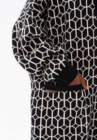 Zwart en crèmekleurig gebreid kledingstuk met geometrisch patroon, voorzien van lange mouwen en een zaklabel "blue shadow." Zachte textuur.