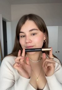 Produit cosmétique noir et doré tenu entre des doigts, avec un design cylindrique. Le modèle porte un collier de perles et un pulls crème.