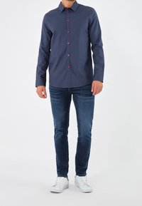 Camicia a maniche lunghe blu navy con bottoni rossi, colletto button-down; abbinata a jeans scuri e sneakers bianche su uno sfondo semplice.
