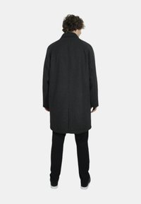 Cappotto nero a motivo houndstooth con taglio dritto, ampio colletto e maniche lunghe, abbinato a pantaloni neri e scarpe bianche, visto da dietro.