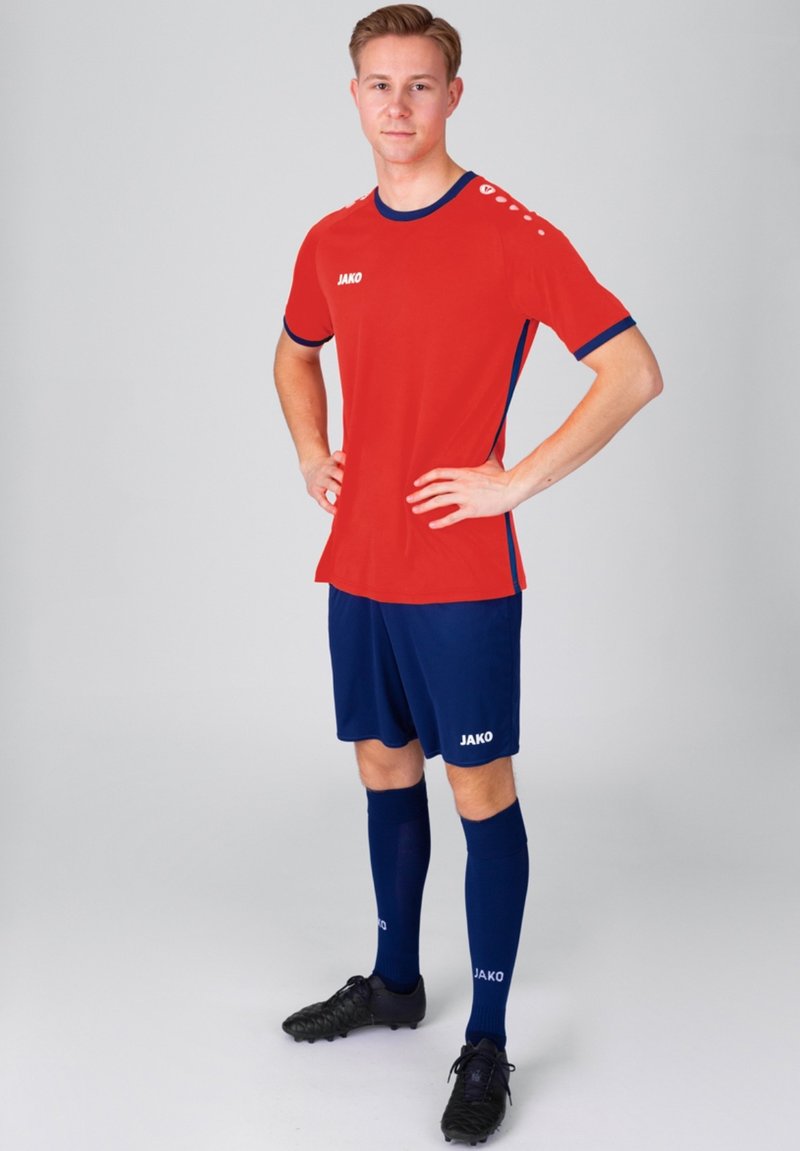 Rotes sportliches Trikot mit blauen Akzenten, kombiniert mit navyfarbenen Shorts und Socken. Enthält ein Logo auf der Brust und den Socken. Strukturiertes Gewebe, figurbetonter Schnitt.