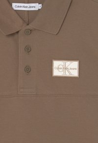 Hnědá polo tričko se třemi knoflíky, štítek Calvin Klein Jeans uvnitř límce a vyšívaná bílá nášivka s nápisem "cK Calvin Klein Jeans" na hrudi.