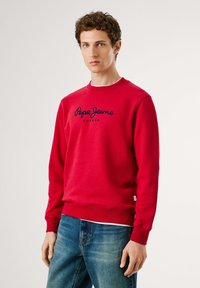 Joven con cabello rizado que lleva una sudadera roja de Pepe Jeans London y jeans azules, de pie contra un fondo claro y liso.